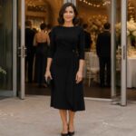 Kobieta w czarnej sukience stoi przed elegancko udekorowaną salą weselną z kwiatami i gośćmi w tle